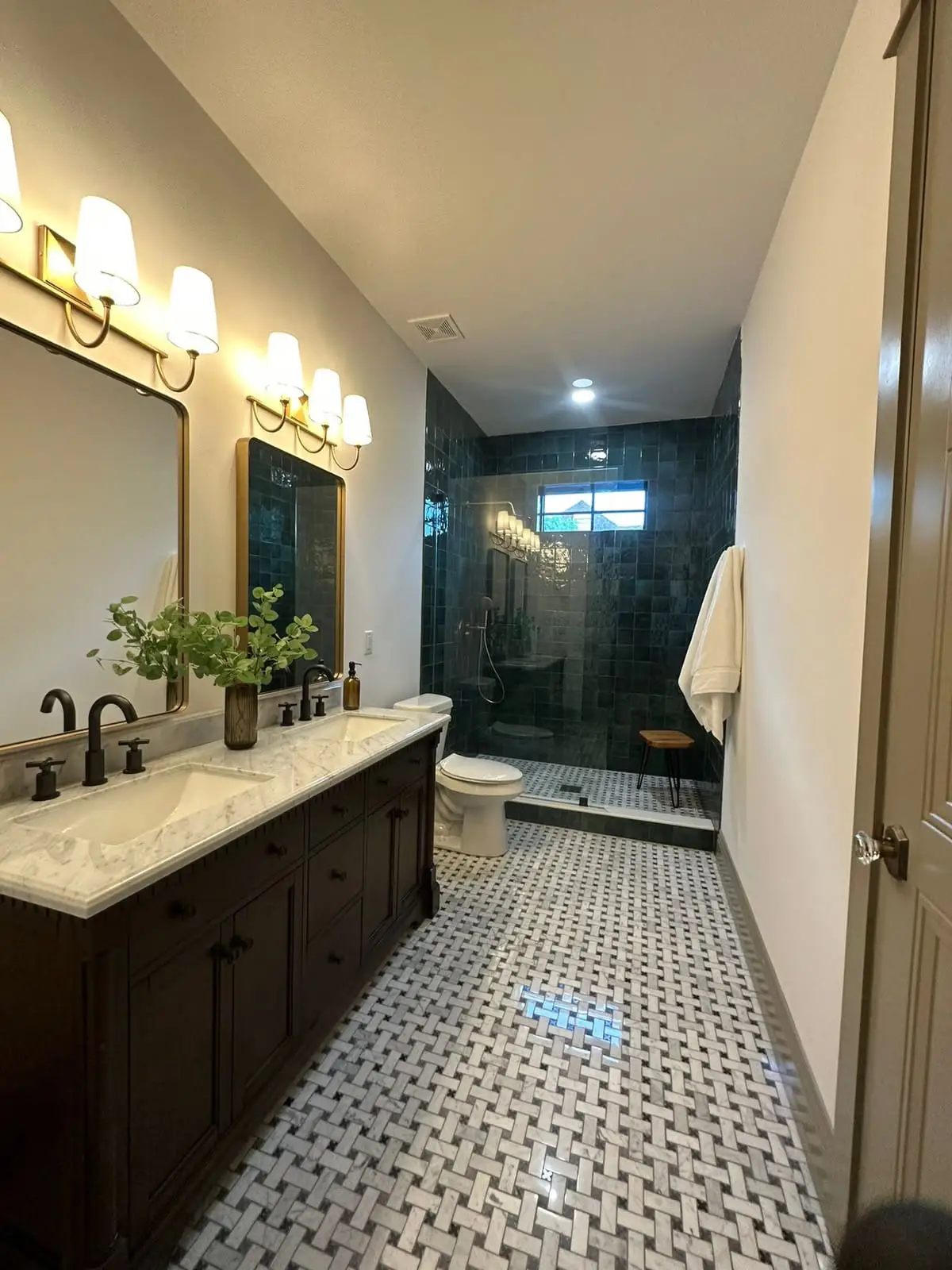 Bathroom with a dark wood double vanity, marble counter, brass sconces and a walk-in shower tiled in deep green, over a basketweave marble floor