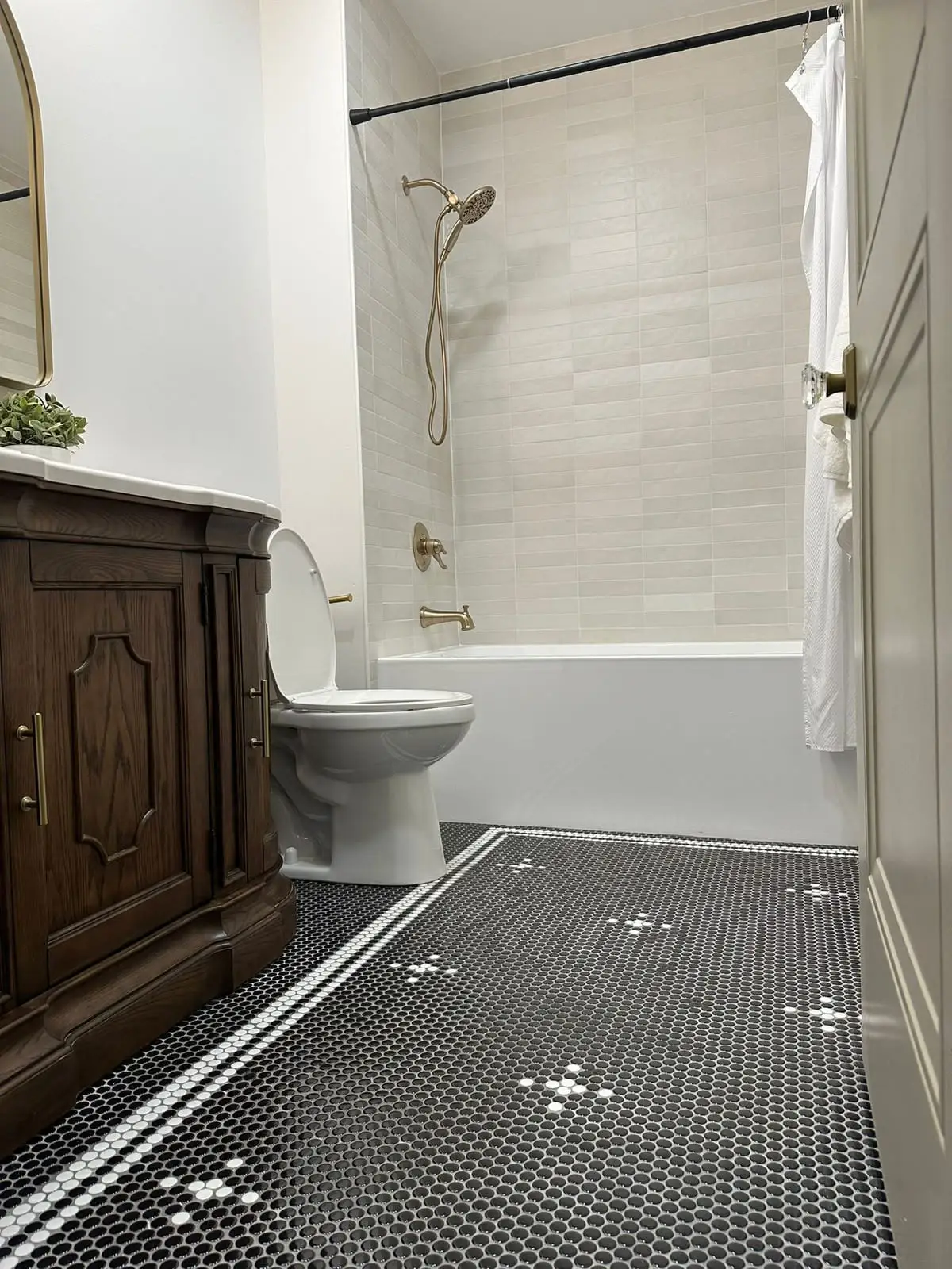 Tub and shower combination in vertically stacked light tile with brass fittings, over a black penny round mosaic floor with a white pattern