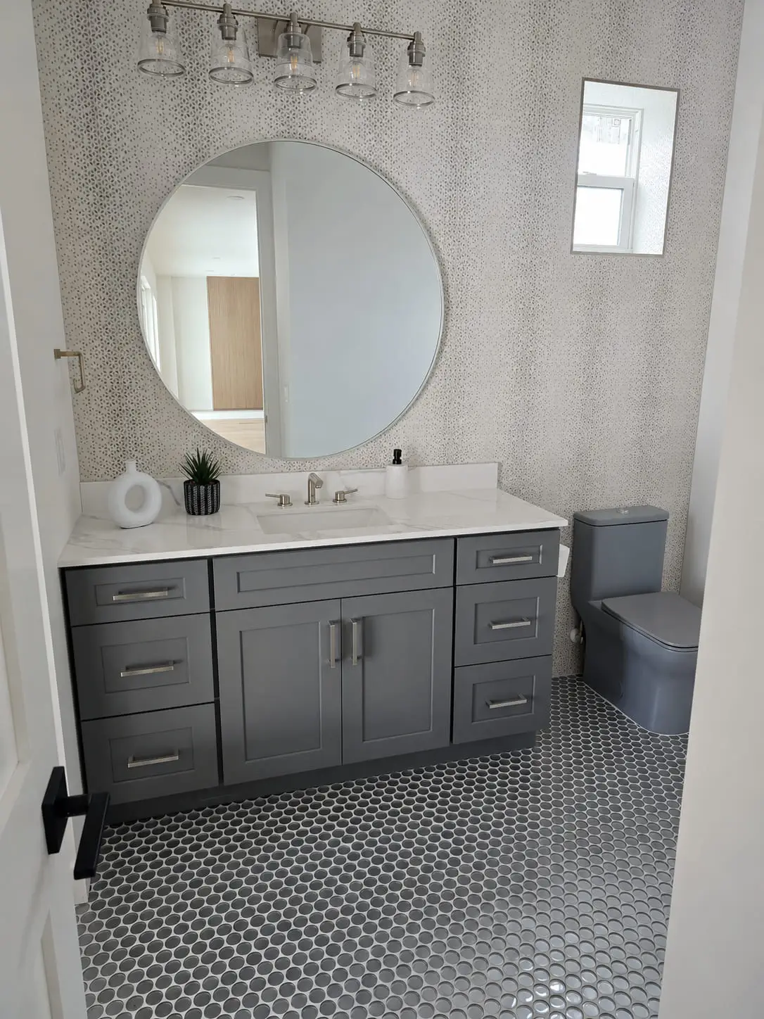 Bathroom remodel with a grey vanity, round mirror and a penny round mosaic floor against a textured feature wall