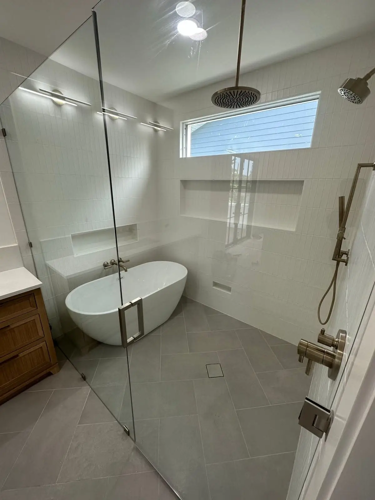 Wet room with a freestanding tub and walk-in shower behind a frameless glass screen, large-format grey floor tile falling to a linear drain and brass fixtures