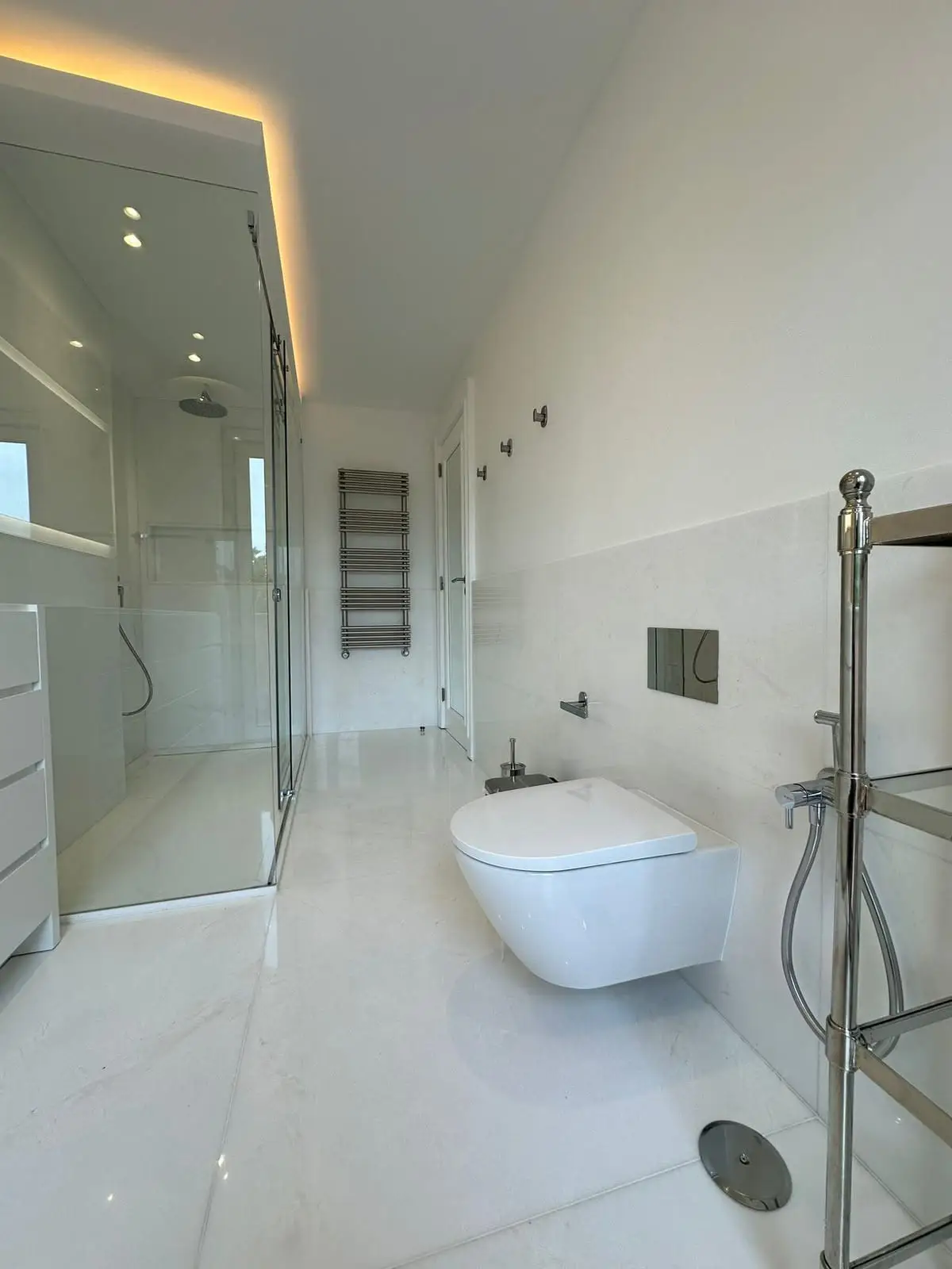 The finished bathroom in white marble-look porcelain, with a glass shower enclosure, wall-hung toilet and a cove-lit ceiling