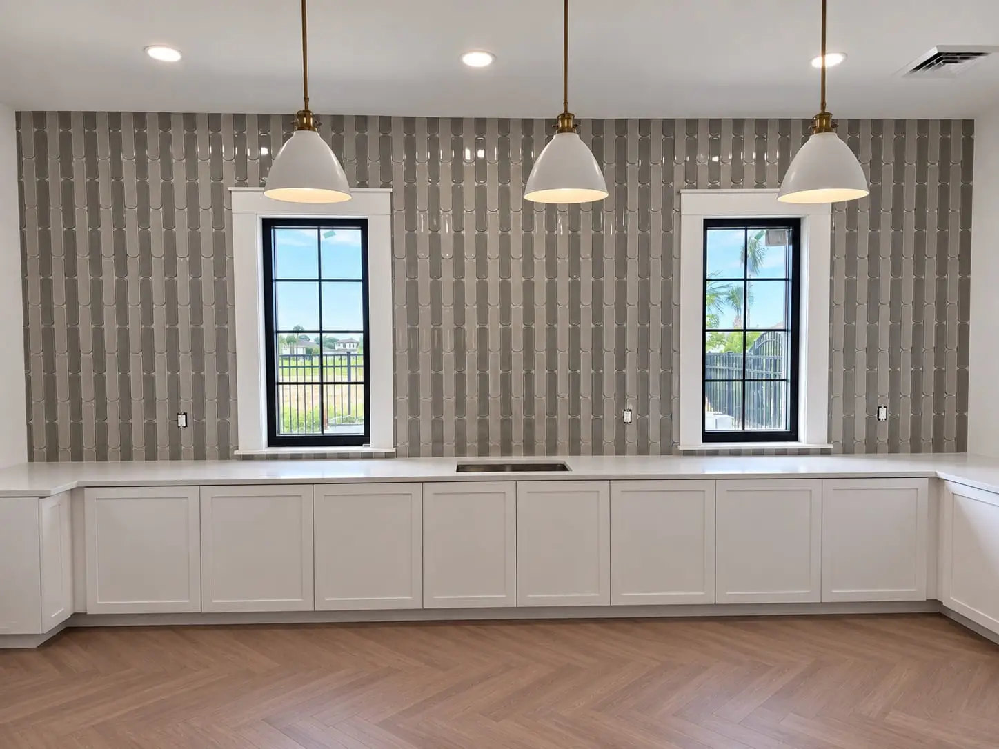 Long counter wall in vertically set grey tile between two windows, under a row of pendant lights