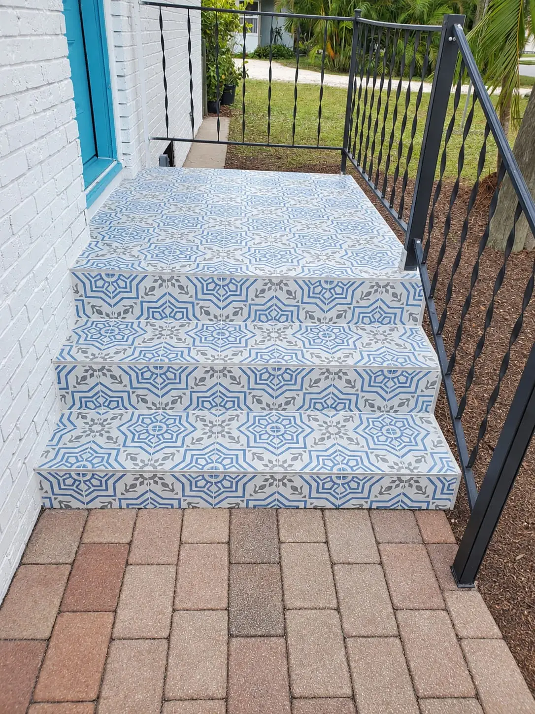 Patterned tile steps meeting a brick paver walkway, with an iron handrail alongside