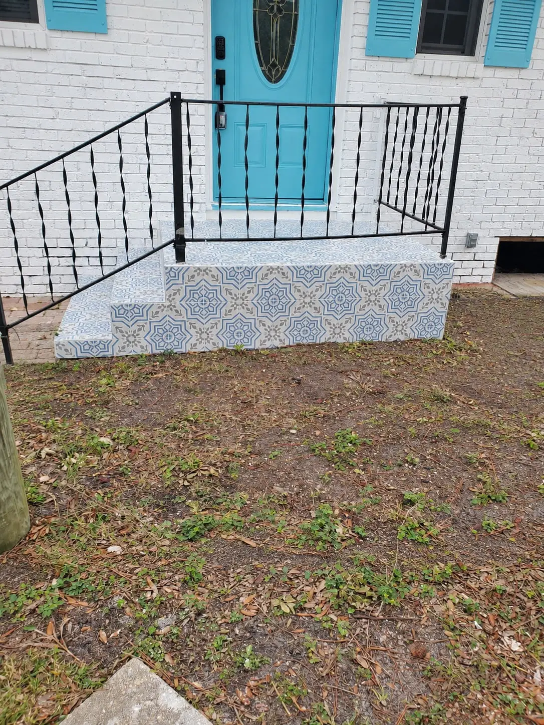 Front steps with blue and white patterned tile risers against a white brick house with a turquoise door and an iron rail